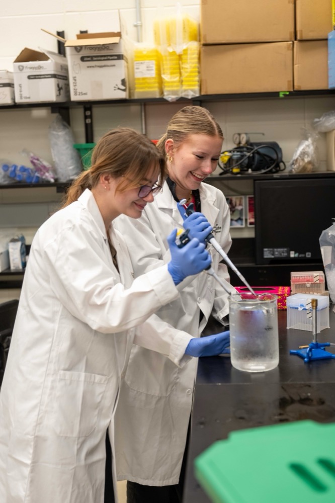 Two Kirkness Scholars in lab coats pipetting a sample in a research lab