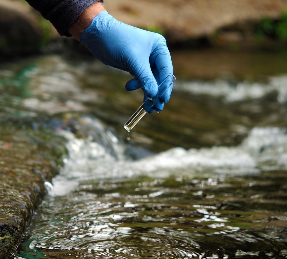 Photo of a water sample being collected from a river. The person collecting the sample is wearing a blue rubber glove and carrying the clear vial.