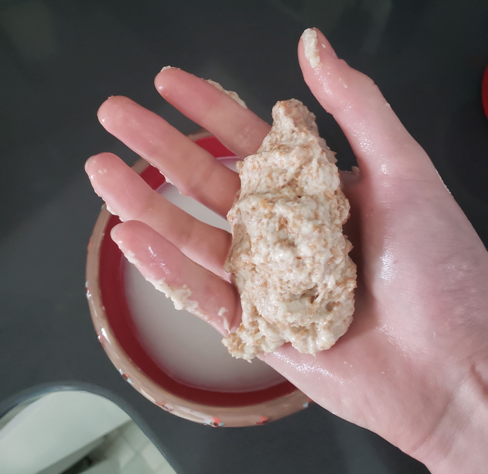 Photo of a hand holding a lump of gluten from flour. The hand is hovering over a bowl with water where the lump of gluten was extracted from.